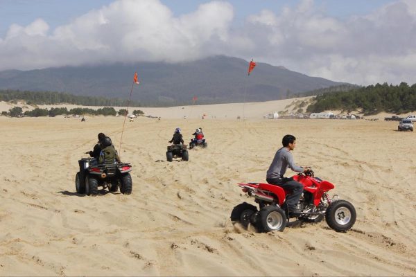 People riding ATVs on wide sandy dunes with grassy hills and a vehicle visible in the background under a hazy sky.