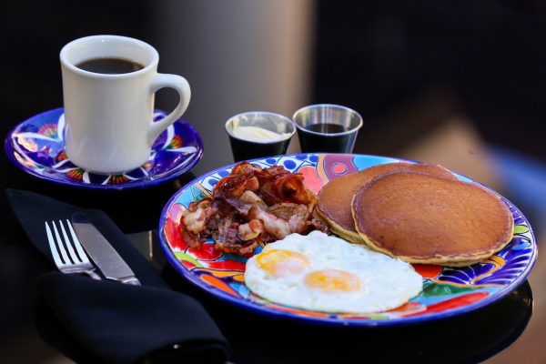 A plate of fried eggs served with bacon, pancakes, and a hot cup of coffee on a brightly colored ceramic plate.