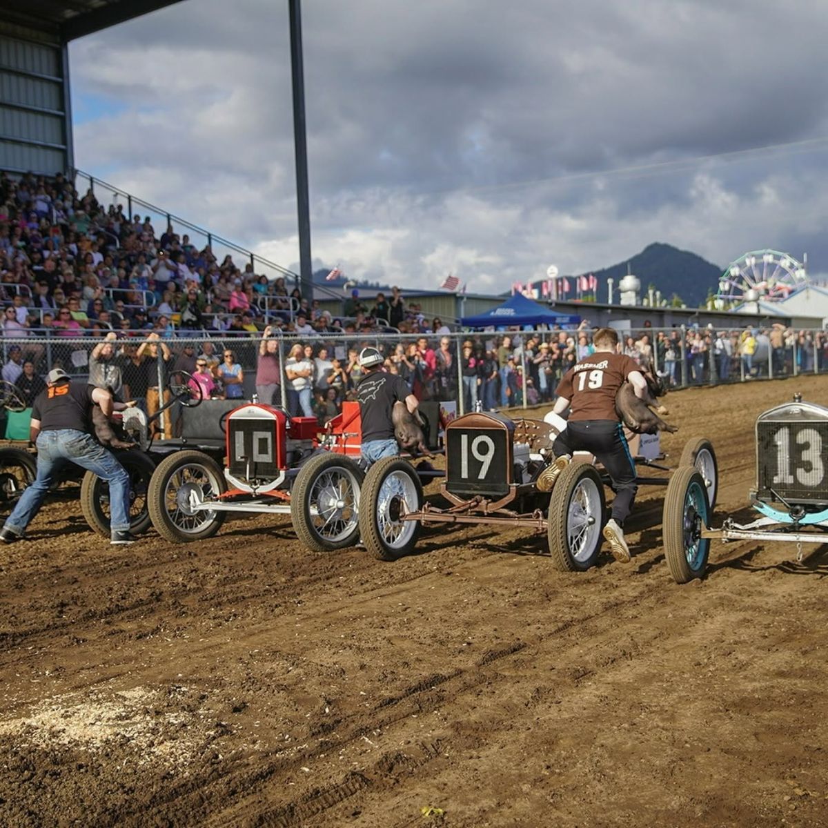 Vintage race cars lined up on a muddy dirt track with a crowded grandstand and distant carnival rides in the background.